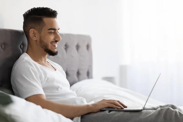 Smiling Young Arab Guy Using Laptop While Relaxing In Bed At Home — Zdjęcie stockowe