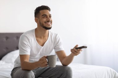 Happy Young Arab Man Watching TV While Relaxing In Bedroom At Home