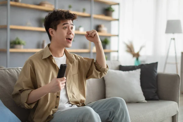 Excited asian guy spending weekend watching tv shaking clenched fist — 图库照片