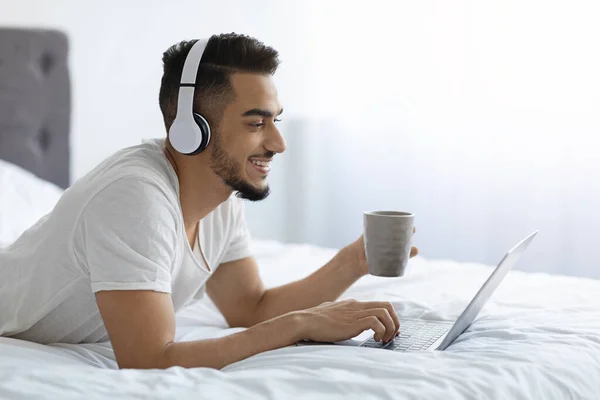 Millennial árabe chico usando auriculares descansando en la cama con el ordenador portátil y café — Foto de Stock