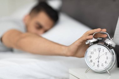 Young Arab Man Reaching Alarm Clock On Bedside Table With Hand