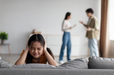 Despair unhappy korean teenage child closes her ears with hands so as not to hear quarrels of young mom and dad