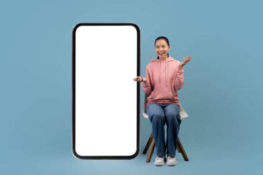 Surprised asian woman sitting on chair by big smartphone, mockup