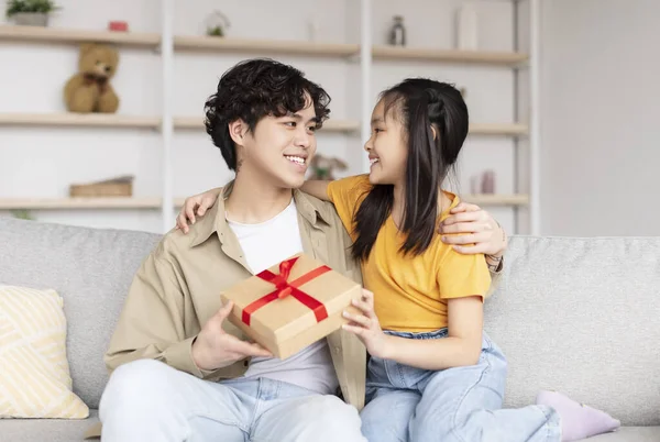Cheerful young asian man hold box with gift from teenager girl sitting on sofa in living room interior — Fotografia de Stock