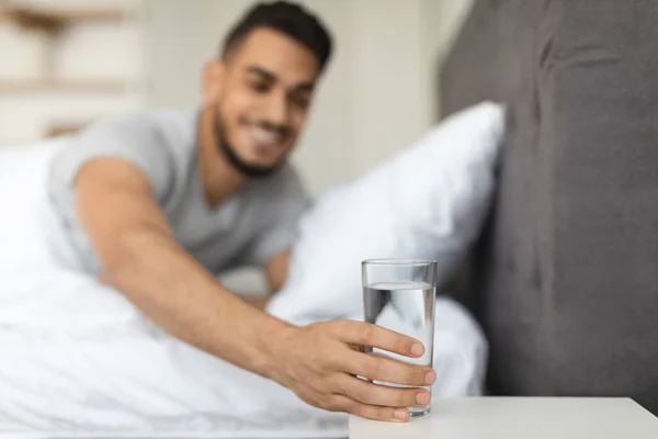 Sonriente hombre de Oriente Medio tomando vaso de agua de la mesita de noche en el dormitorio —  Fotos de Stock