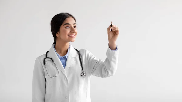 Cheerful millennial cute indian lady medical worker in uniform writes on virtual board in air — Stock Photo, Image