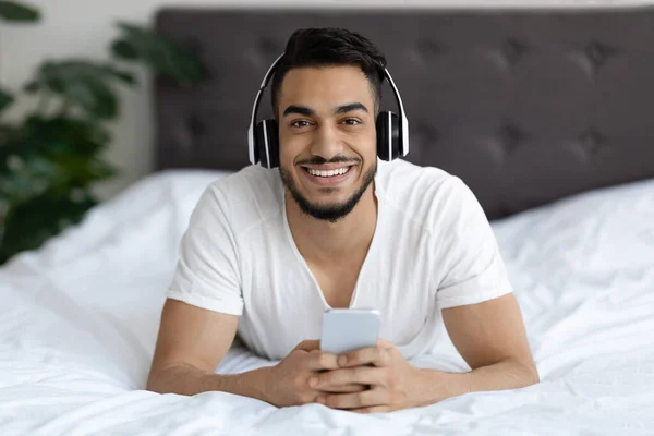 Happy Young Arab Guy Wearing Wireless Headphones Resting In Bed With Smartphone — Stockfoto