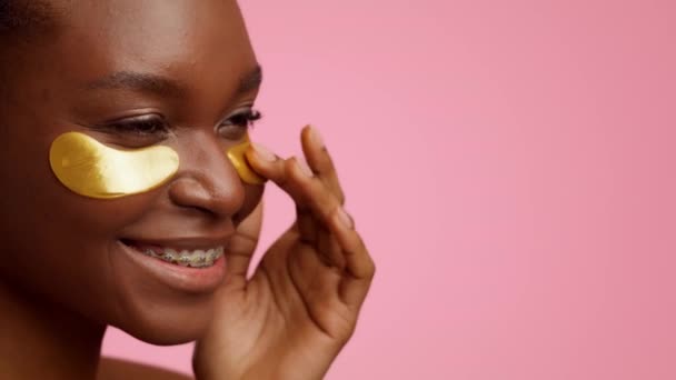 Black Woman Applying Golden Patches Under Eyes, Pink Background, Cropped — Vídeo de Stock