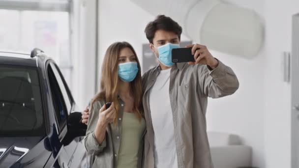 Young happy man and woman wearing protective medical masks bought new car, recording video on smartphone with keys — Video