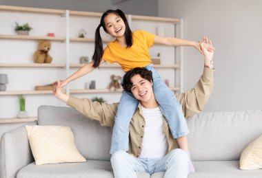 Cheerful young asian man holds on his shoulders teenager girl, sit on sofa in living room interior