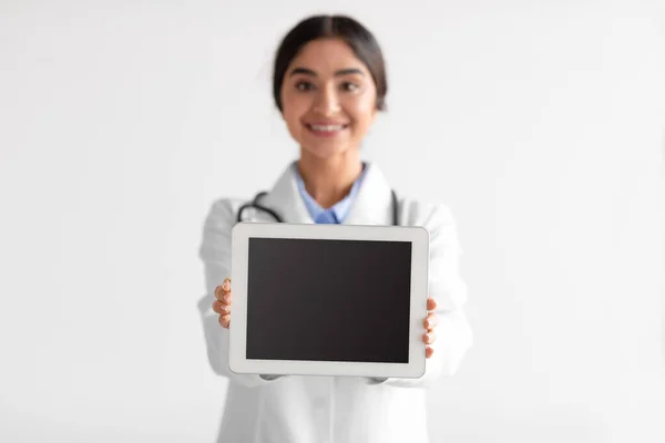 Smiling millennial hindu woman nurse or doctor in uniform shows digital tablet with empty screen — 스톡 사진