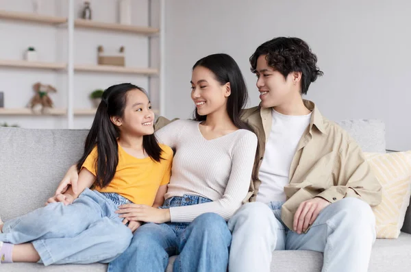 Happy handsome millennial asian couple watching at teen daughter, hugging, sitting on sofa in living room — Stockfoto