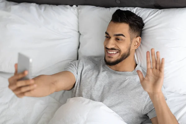 Comunicación virtual. Chico árabe feliz haciendo videollamada con Smartphone en la cama — Foto de Stock