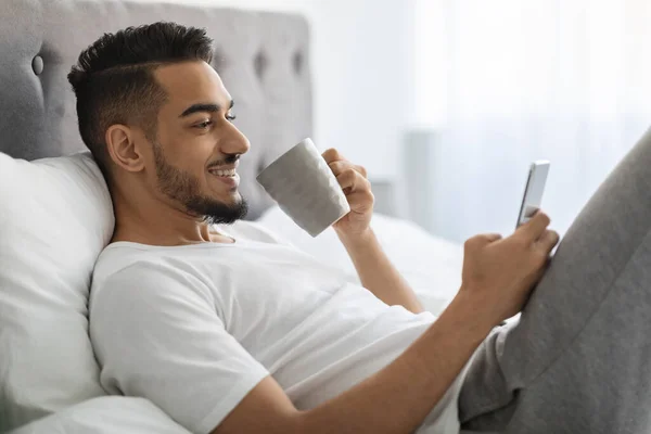 Χαρούμενος νεαρός Άραβας χαλαρώνοντας στο κρεβάτι με Smartphone και καφέ — Φωτογραφία Αρχείου