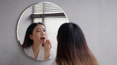 Lip Care. Beautiful asian woman applying moisturising chapstick on lips in bathroom