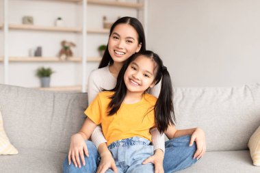 Glad smiling young korean female hugging teen girl sitting on sofa in living room interior