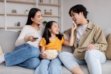 Cheerful young asian lady, man and teenager girl watch tv, daughter feeding daddy popcorn, sits on sofa