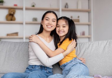 Satisfied pretty millennial chinese woman and teenage girl hugging, sitting on sofa in living room