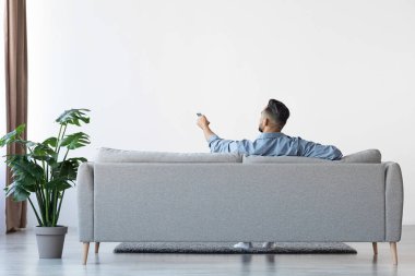Man Sitting On Couch Opening Air Conditioner With Remote Controller, Rear View