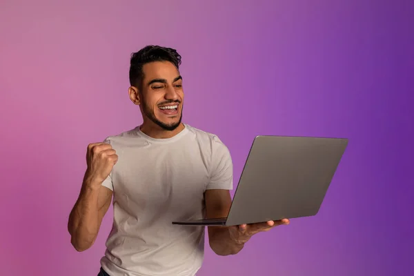 Cool young Arab man holding laptop, making YES gesture, excited over huge online lottery win or success in neon light — Fotografia de Stock