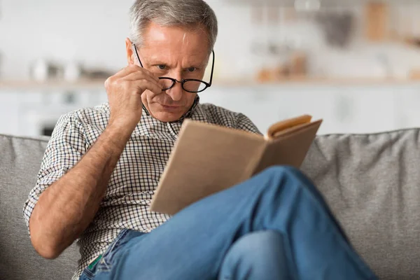 Senior Man With Poor Eyesight Reading Book Above Eyeglasses Indoor — Stockfoto
