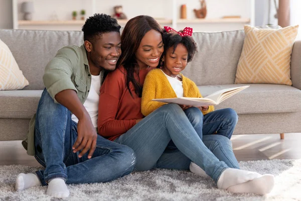 Alegre familia afroamericana con la pequeña hija leyendo el libro juntos en casa — Foto de Stock