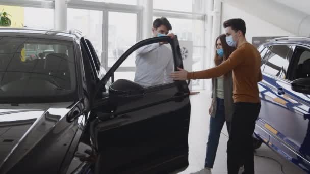 Los clientes casados jóvenes de la pareja que buscan el automóvil nuevo en el salón, distribuidor profesional del hombre que muestra el vehículo a la mujer — Vídeos de Stock