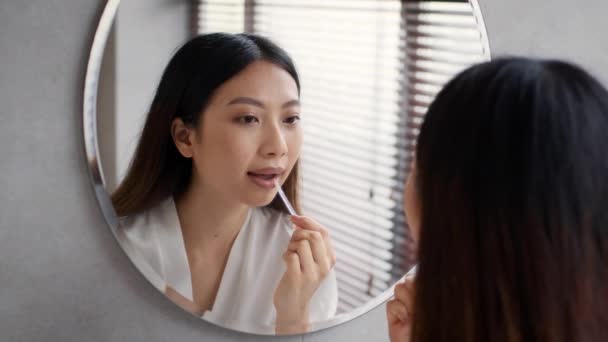 Rutina de maquillaje. Hermosa mujer asiática aplicando brillo de labios cerca del espejo en el baño — Vídeos de Stock