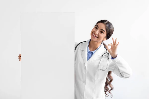 Glad young cute indian lady doctor in uniform shows ok sign with hand holds big banner with blank space — Foto Stock