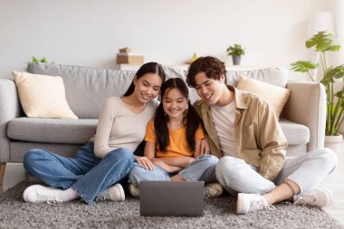 Satisfied millennial japanese mom, dad and teen daughter sitting on floor and watching video on laptop