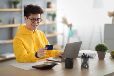 Smiling asian guy showing using credit card at home