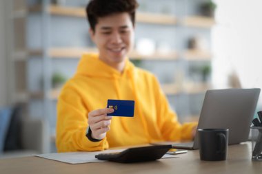 Smiling asian guy showing using credit card at home