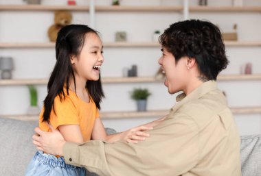 Happy asian millennial man and teen daughter look at each other with open mouths, have fun