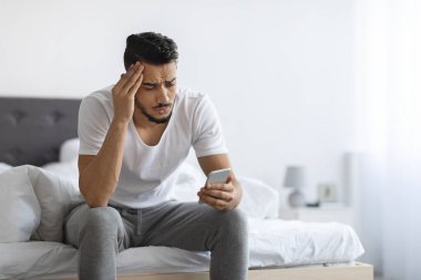 Bad News. Upset Arab Man Sitting On Bed And Looking At Smartphone