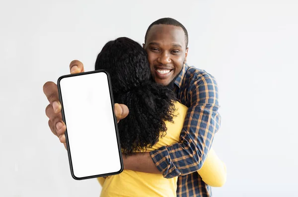 Handsome black guy hugging his girlfriend and showing smartphone, mockup — стоковое фото