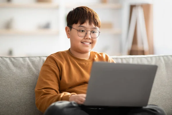 Cheerful chinese kid sitting on couch with laptop at home — Zdjęcie stockowe