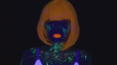 Young asian woman wearing orange wig with fluorescent makeup coming to camera and dancing over black background