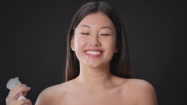 Skin moisturizing concept. Young playful asian woman spraying water on her face and laughing to camera, black background — Stockvideo