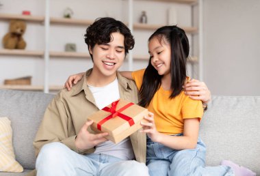 Smiling happy chinese young guy receives gift from teenager daughter in room interior
