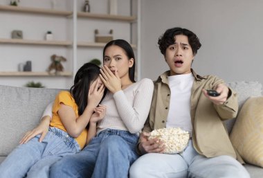 Frightened shocked asian millennial wife, husband and adolescent daughter watching scary movie and eating popcorn