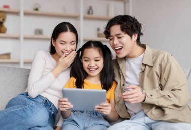 Laughing young asian husband, wife and teen daughter watching funny video on tablet, surfing in internet