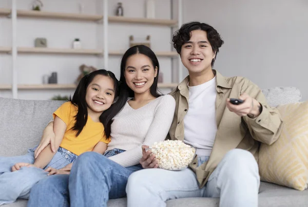 Happy young chinese wife, husband and teenage girl watching tv, eating popcorn, sitting on sofa in living room interior — ストック写真