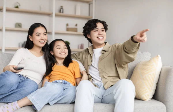 Smiling japanese young man shows on empty space with hand to wife and teen girl, sit on sofa in living room — Stockfoto