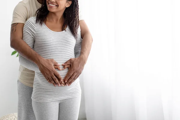 Улыбающийся молодой африканский американец обнимает беременную женщину животом, делает сердце руками в интерьере комнаты, неузнаваемым — стоковое фото