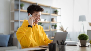 Neşeli Asyalı adam bilgisayar kullanarak cep telefonuyla konuşuyor ve çalışıyor.