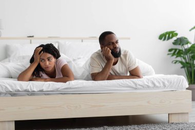 Offended sad unhappy young african american husband ignores wife, lie on comfortable bed in bedroom interior