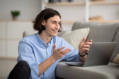 Young guy sitting near sofa in front of laptop, gesturing and smiling, having video call with friends or colleagues