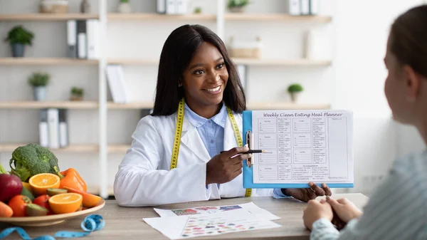 Consulta com nutricionista. Jovem dietologista negra mostrando exemplo paciente de menu semanal — Fotografia de Stock