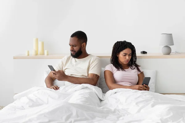 Offended sad young african american guy ignores lady and typing on smartphones on comfortable bed — 스톡 사진