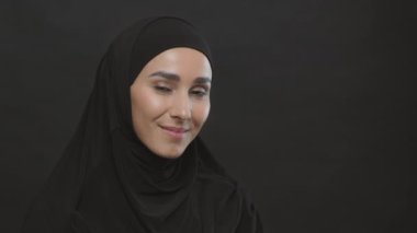 Siyah tesettürlü mutlu Müslüman kadın kameraya gülümsüyor, siyah stüdyo arka planında boş alan ile poz veriyor.